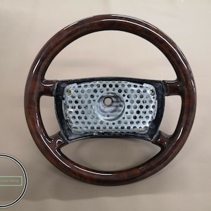 Pode incluir: Um volante de madeira castanho escuro com um cubo central preto e uma grelha de malha metálica prateada. O volante tem um design clássico e está em bom estado.