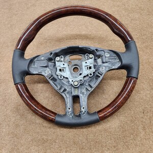 May include: A brown wood steering wheel with a black leather rim. The steering wheel has a silver metal centre with a black plastic spoke.