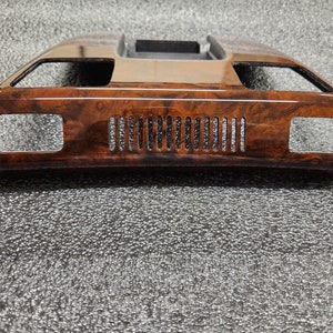 May include: A car interior trim piece with a wood grain effect. The trim is rectangular with two side openings and a series of vertical slots in the centre. The top section has a shiny, metallic finish.