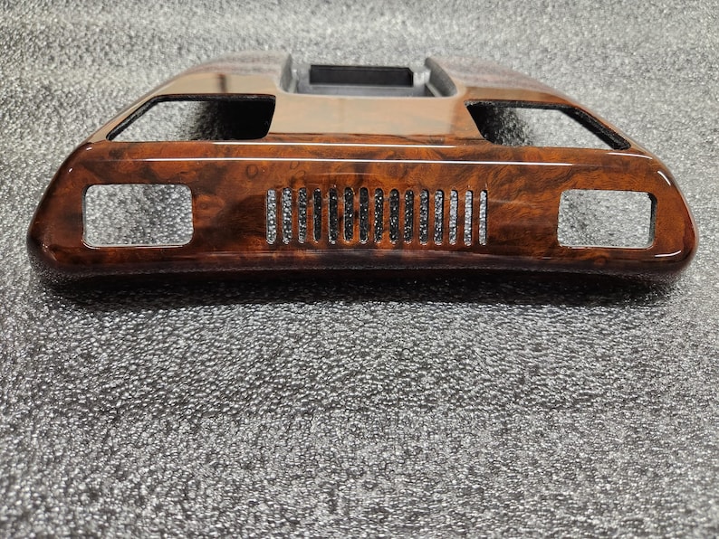 May include: A car interior trim piece with a wood grain effect. The trim is rectangular with two side openings and a series of vertical slots in the centre. The top section has a shiny, metallic finish.