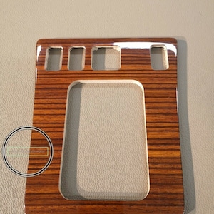 以下が含まれることがあります： 長方形の切り抜きと5つの小さな長方形の切り抜きがある、茶色の木目調の車の内装トリム。