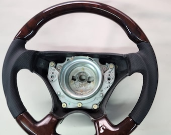 May include: A car steering wheel with a dark wood grain finish and black leather grip sections. The wheel has a metal center with mounting hardware and a small logo on the lower right spoke.