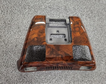 May include: A car dashboard trim piece with a glossy, wood-grain finish. The trim is a dark brown color with lighter brown swirls and patterns. It has a rectangular shape with a central opening and two side openings.