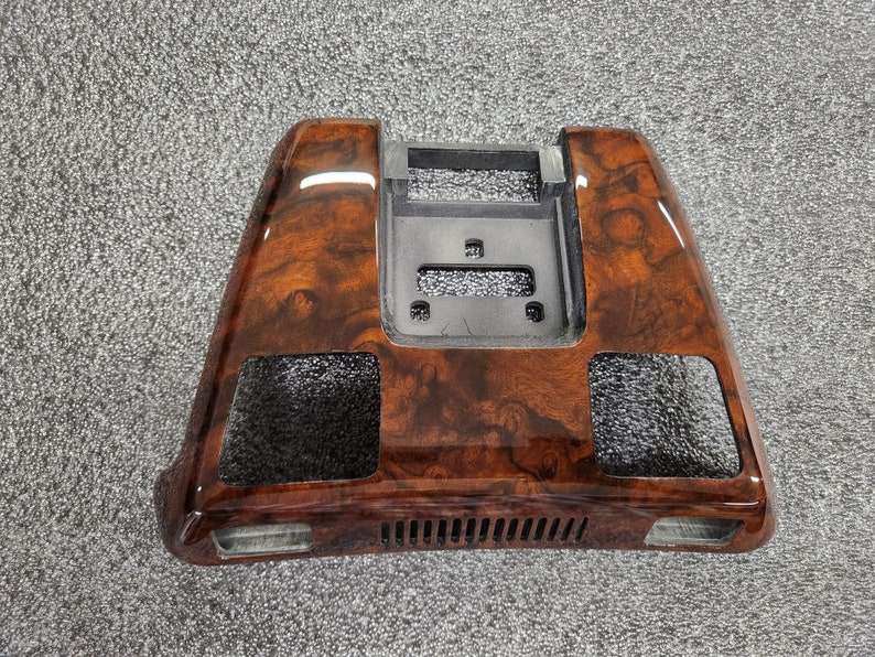 May include: A car dashboard trim piece with a glossy, wood-grain finish. The trim is a dark brown colour with lighter brown swirls and patterns. It has a rectangular shape with a central opening and two side openings.
