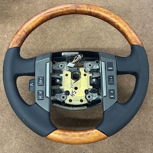 Può includere: Un volante in legno marrone e pelle nera con comandi argentati. I comandi includono pulsanti per "Modalità", "+", "-" e "< >".