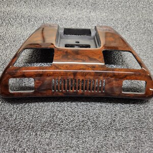 May include: A wood-effect car dashboard trim piece. The trim is a dark brown colour with a glossy finish. It features rectangular cutouts and a series of small, vertical vents. The piece has a curved, angular design.