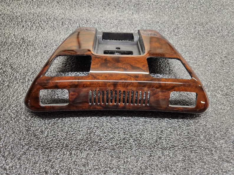May include: A wood-effect car dashboard trim piece. The trim is a dark brown colour with a glossy finish. It features rectangular cutouts and a series of small, vertical vents. The piece has a curved, angular design.