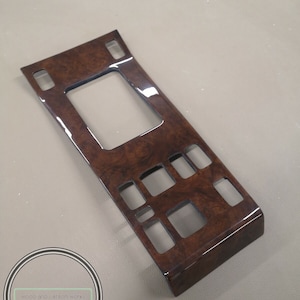 以下が含まれることがあります： 光沢のある仕上げと複数の長方形の切り抜きが施された、茶色の木製の車のインテリアトリムパネル。パネルは右側に湾曲したエッジがあります。