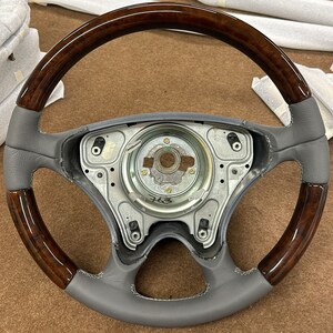 Könnte beinhalten: Ein graues Lederlenkrad mit einem Holzrand. Das Lenkrad hat eine silberne Mitte mit dem Wort "Airbag" darauf gedruckt. Das Lenkrad ist in gutem Zustand.