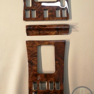 Può includere: Un set di tre pezzi di rivestimento interno per auto in legno. I pezzi sono marroni con una trama a legno e hanno una finitura lucida. I pezzi sono rettangolari e hanno una varietà di ritagli per pulsanti e interruttori.