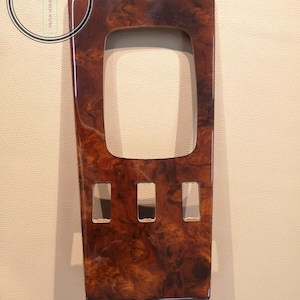 Puede incluir: Una moldura interior de coche de madera marrón con tres recortes rectangulares. La moldura tiene un acabado brillante.