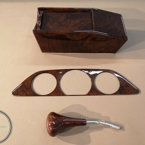 May include: A set of three brown wood car interior trim pieces. The pieces include a rectangular box, a long rectangular panel with three circular cutouts, and a brown wood gear shift knob with a silver metal lever.