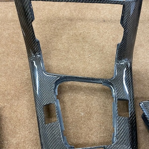 Puede incluir: Una pieza de moldura interior de coche de fibra de carbono negra con un recorte rectangular en el centro. La pieza de moldura tiene una superficie texturizada y un acabado brillante.