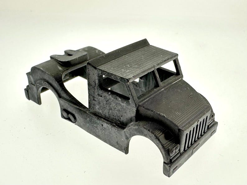 Pode incluir: Uma cabine de caminh&atilde;o modelo de pl&aacute;stico preto com uma grelha frontal detalhada e um teto elevado. A cabine n&atilde;o tem ca&ccedil;amba nem rodas.