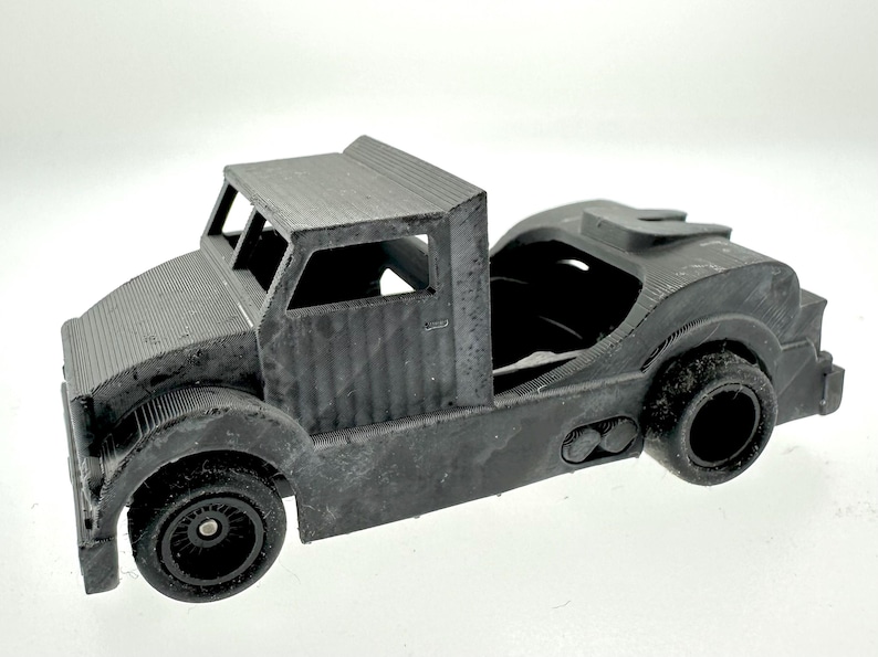 Pode incluir: Um caminh&atilde;o de brinquedo de pl&aacute;stico preto com um reboque de plataforma. O caminh&atilde;o tem uma cabine detalhada com um p&aacute;ra-brisa e um teto. O reboque est&aacute; vazio e tem um engate na parte traseira.