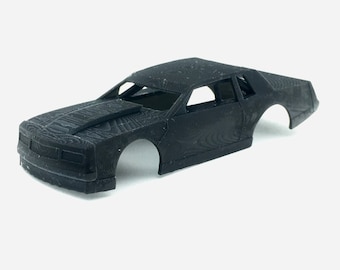 Pode incluir: Uma carroceria de carro modelo de plástico preto, provavelmente para um modelo de corrida de stock car. A carroceria não está pintada e tem uma superfície texturizada.
