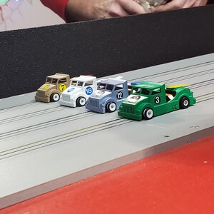 Pode incluir: Quatro carros de corrida de brinquedo alinhados em uma pista. Os carros s&atilde;o verdes, azuis, brancos e marrons. Os carros t&ecirc;m n&uacute;meros: 3, 12, 18 e 7.