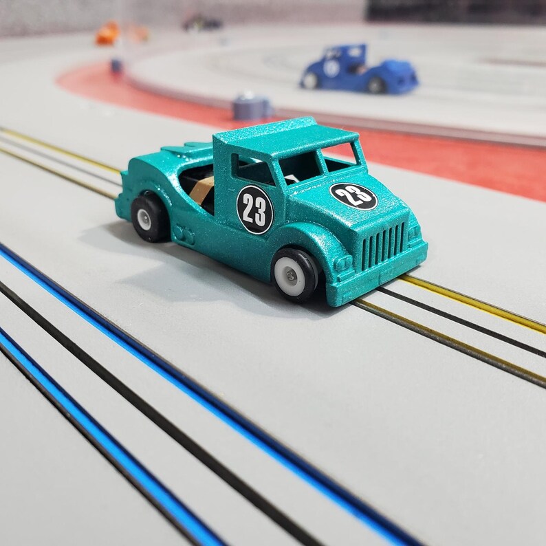 Pode incluir: Um caminh&atilde;o de brinquedo azul turquesa com o n&uacute;mero 23 preto de cada lado. O caminh&atilde;o est&aacute; em uma pista cinza com linhas pretas e azuis.