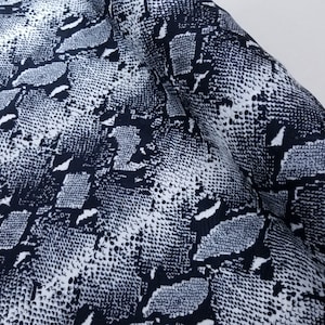 Puede incluir: Primer plano de una tela con estampado de piel de serpiente. El diseño presenta un patrón repetitivo de escamas grises oscuras, grises claras y blancas. La tela está doblada, creando sombras y resaltando la textura del material.