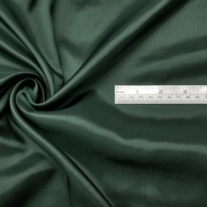 May include: A close-up of a deep green satin fabric. The fabric is smooth and silky, with a slight sheen. A ruler is placed on top of the fabric, showing the fabric's texture.