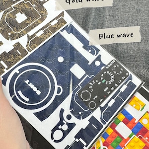 May include: Three camera skin designs: a gold wave pattern, a blue wave pattern, and a colourful toy brick pattern. The designs are printed on white paper and are ready to be applied to a camera.