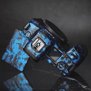 Puede incluir: Una c&aacute;mara Canon EOS con un dise&ntilde;o de camuflaje azul y negro. La c&aacute;mara tiene una lente negra y una peque&ntilde;a pantalla. La c&aacute;mara est&aacute; sobre una superficie reflectante.