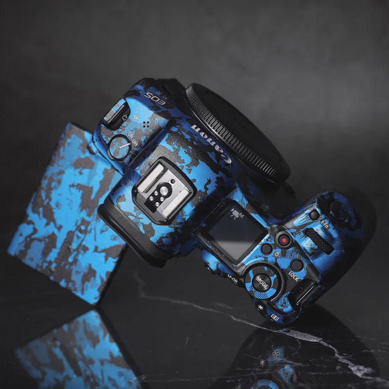 Puede incluir: Una c&aacute;mara Canon EOS con un dise&ntilde;o de camuflaje azul y negro. La c&aacute;mara tiene una lente negra y una peque&ntilde;a pantalla. La c&aacute;mara est&aacute; sobre una superficie reflectante.