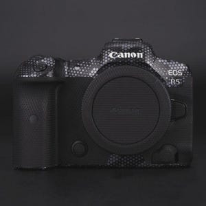Puede incluir: Una c&aacute;mara Canon EOS R5 negra con un dise&ntilde;o estampado. La c&aacute;mara tiene una tapa de lente y el logotipo de Canon es visible. La c&aacute;mara est&aacute; sobre una superficie negra.