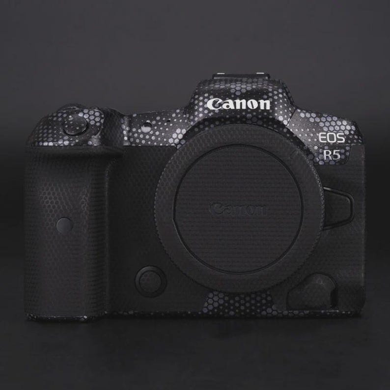 Puede incluir: Una c&aacute;mara Canon EOS R5 negra con un dise&ntilde;o estampado. La c&aacute;mara tiene una tapa de lente y el logotipo de Canon es visible. La c&aacute;mara est&aacute; sobre una superficie negra.