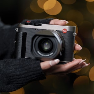 Puede incluir: Una cámara Leica plateada y negra con un logotipo rojo. La cámara tiene una empuñadura de cuero negro y una lente negra.