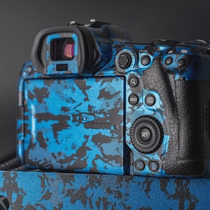 Puede incluir: Una c&aacute;mara Canon con un dise&ntilde;o de camuflaje azul y negro. La parte trasera de la c&aacute;mara tiene botones para informaci&oacute;n, configuraci&oacute;n y revisi&oacute;n de im&aacute;genes. El logotipo de Canon es visible.