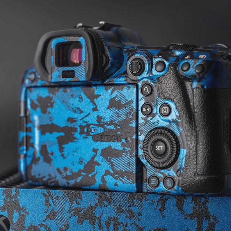 Puede incluir: Una c&aacute;mara Canon con un dise&ntilde;o de camuflaje azul y negro. La parte trasera de la c&aacute;mara tiene botones para informaci&oacute;n, configuraci&oacute;n y revisi&oacute;n de im&aacute;genes. El logotipo de Canon es visible.