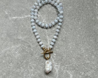 Puede incluir: Un collar de cuentas azul claro con un cierre dorado y un gran colgante de perla blanca irregular. El collar se muestra sobre una superficie gris, mostrando el diseño y el contraste de color.
