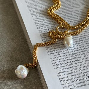 May include: A gold chain necklace with a large, irregular, white pearl pendant. The necklace is layered and has a chunky, textured design. The pearl is hanging from a small gold ring.