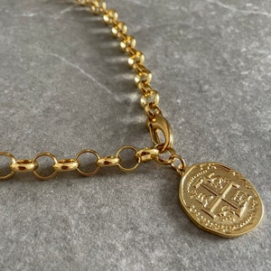 May include: A gold chain necklace with a large, round pendant featuring an intricate design. The pendant has a raised, textured surface with a crest-like image.