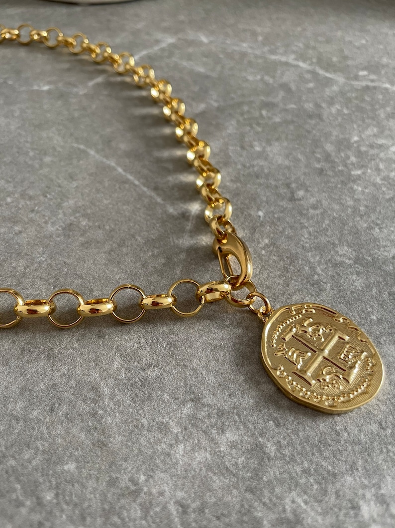 May include: A gold chain necklace with a large, round pendant featuring an intricate design. The pendant has a raised, textured surface with a crest-like image.
