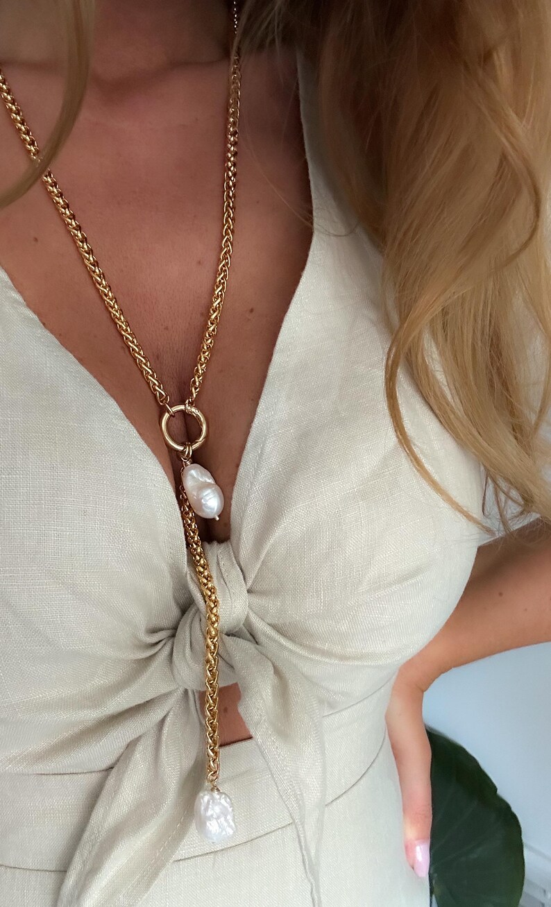 May include: A gold chain necklace with a large pearl pendant. The necklace is knotted at the chest and has a second pearl pendant at the bottom.