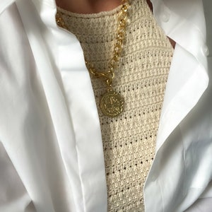 May include: A gold chain necklace with a large, round pendant featuring a detailed design. The necklace is worn over a white crochet top and a white button-down shirt.
