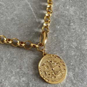 May include: A gold chain necklace with a large, round, gold coin pendant featuring an intricate design. The coin has a raised, textured cross in the center.