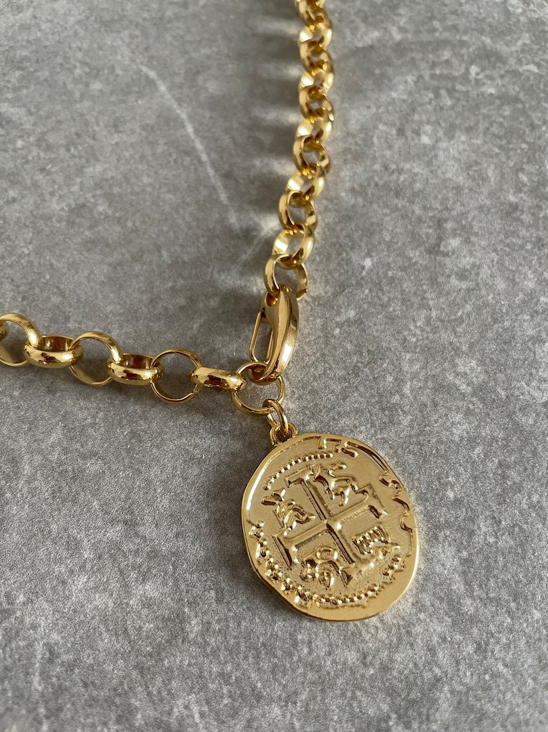 May include: A gold chain necklace with a large, round, gold coin pendant featuring an intricate design. The coin has a raised, textured cross in the center.