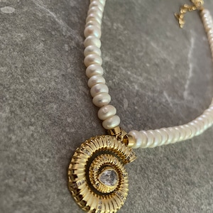 May include: A white pearl necklace with a gold spiral pendant featuring a clear rhinestone. The pendant is in the shape of a nautilus shell.