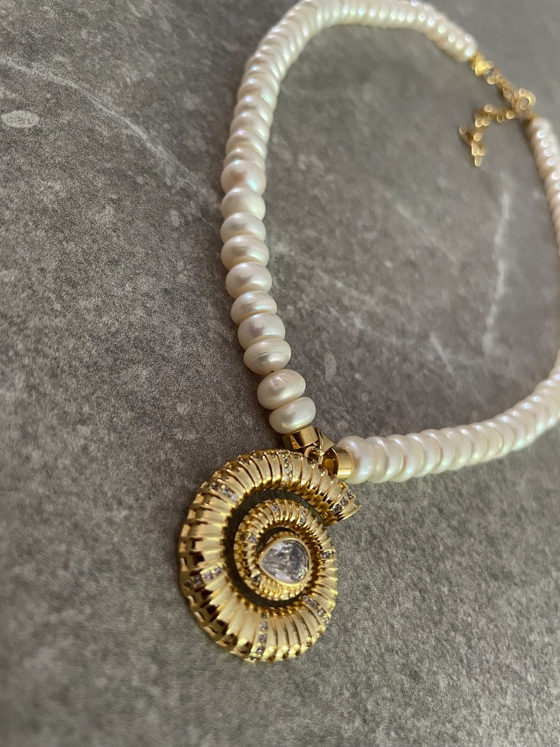 May include: A white pearl necklace with a gold spiral pendant featuring a clear rhinestone. The pendant is in the shape of a nautilus shell.