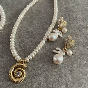 May include: A pearl necklace with a gold seashell pendant and a pair of pearl and crystal earrings. The necklace and earrings are on a gray background.