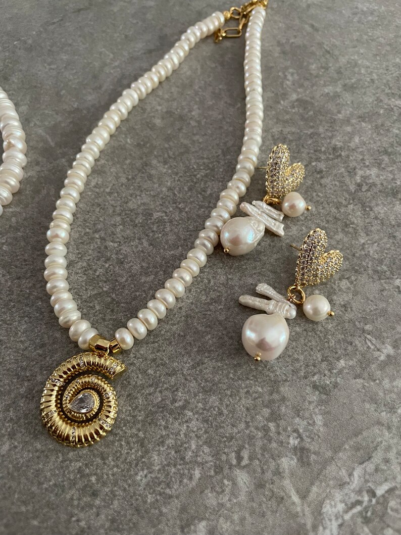 May include: A pearl necklace with a gold seashell pendant and a pair of pearl and crystal earrings. The necklace and earrings are on a gray background.