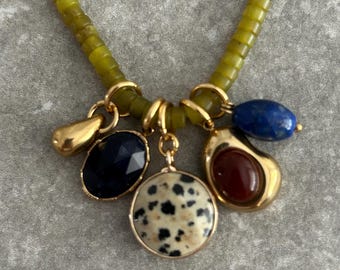 Op de afbeelding: Een ketting met een groene kralenketting en goudkleurige accenten. De ketting heeft verschillende bedels, waaronder een gouden druppel, een donkerblauwe ovale steen, een ronde steen met zwarte stippen en een roodbruine steen.