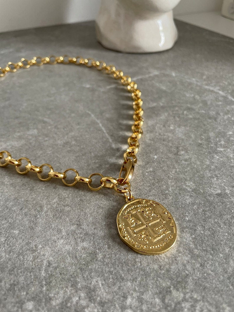 May include: A gold chain necklace with a large, round, gold coin pendant. The coin has an intricate design with a cross in the center.