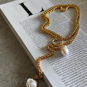 May include: A gold chain necklace with a large, irregular pearl pendant. The chain is made of two interwoven strands, creating a textured look. The pearl is off-white and has a slightly iridescent sheen.