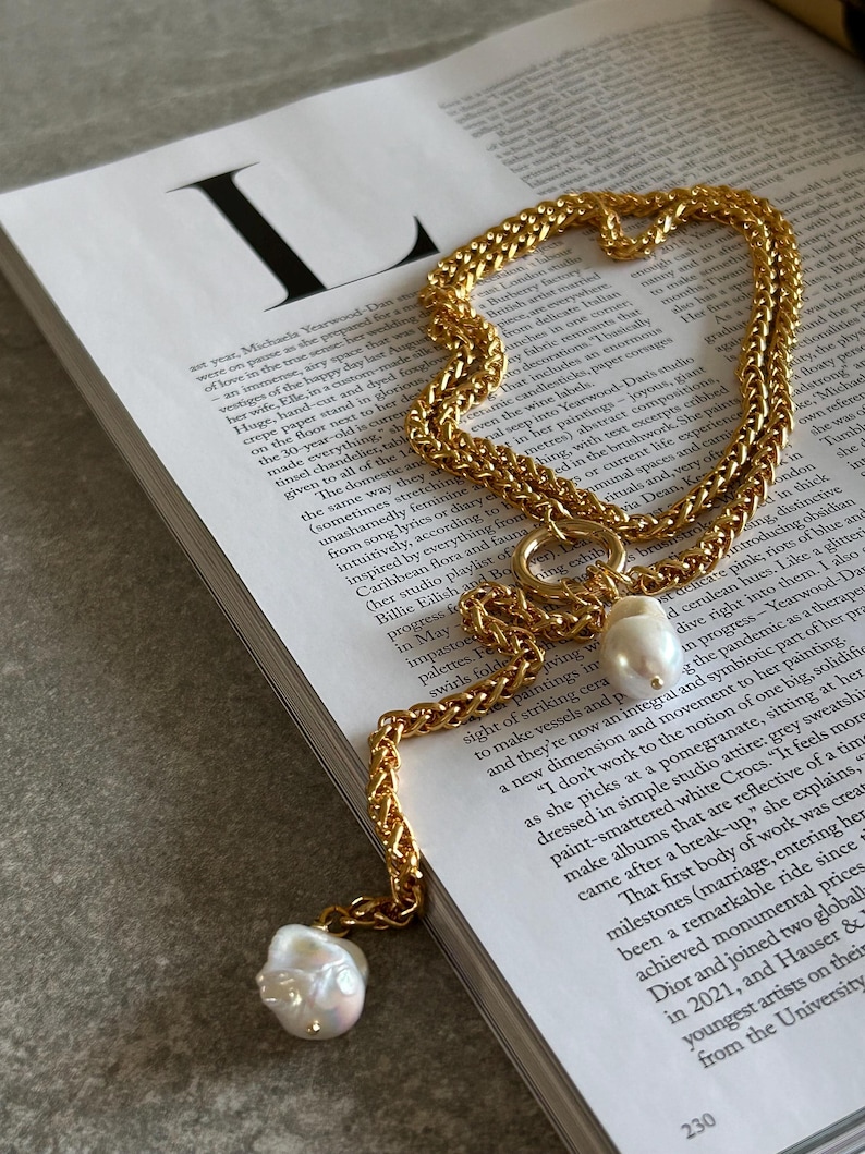 May include: A gold chain necklace with a large, irregular pearl pendant. The chain is made of two interwoven strands, creating a textured look. The pearl is off-white and has a slightly iridescent sheen.