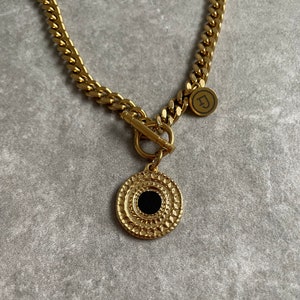 May include: A gold chain necklace with a large round pendant featuring a black centre and intricate gold detailing. The chain has a toggle clasp and a small gold charm with the letter "D" on it.
