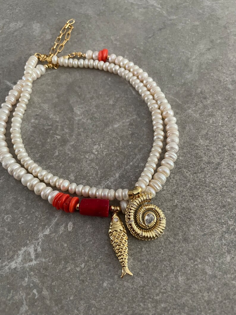 May include: A double strand pearl necklace with a red coral bead and gold charms, including a fish and a spiral shell with a clear rhinestone.